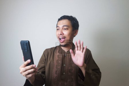 Fröhlicher junger asiatischer Muslim, der ein Mobiltelefon benutzt und isoliert vor weißem Hintergrund wegschaut
