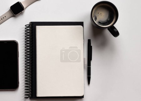 Foto de Moderna mesa de escritorio de oficina blanca con cuadernos, bolígrafo, smartphone, reloj inteligente y taza de café. Página de cuaderno en blanco para la entrada de texto en el medio. Vista superior, plano. - Imagen libre de derechos