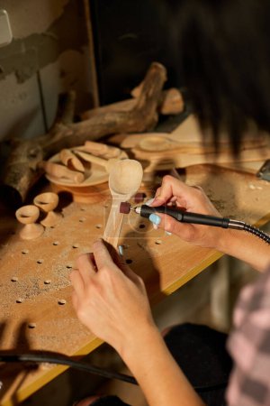 Téléchargez les photos : Femme utilisant des outils de travail de puissance graveur pour ustensiles en bois cuillère, sculpture, rectifieuse lors de l'artisanat, Création de souvenirs artisanaux, femme dans l'atelier. - en image libre de droit