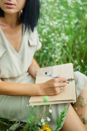 Frau mit Stift schreiben oder malen, Handschrift auf Notizbuch auf blumenblühender Wiese, auf dem Land im Freien, auf dem Feld an Sommertagen auf der Wiese, die Natur spüren, entspannen, allein reisen.
