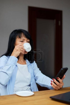 Foto de Anciana mujer de negocios relajarse en el lugar de trabajo, el uso de teléfono móvil y beber café mientras toma un descanso durante las horas de trabajo en la oficina. Descanso femenino en el trabajo - Imagen libre de derechos