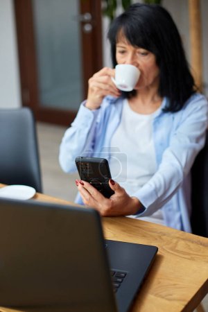 Foto de Anciana mujer de negocios relajarse en el lugar de trabajo, el uso de teléfono móvil y beber café mientras toma un descanso durante las horas de trabajo en la oficina. Descanso femenino en el trabajo - Imagen libre de derechos