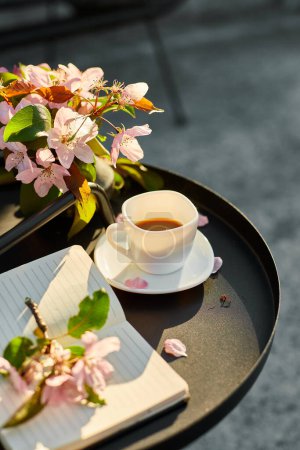 Taza de flores de café y cuaderno en la pequeña mesa negra en la terraza en casa en un día soleado, espacio de trabajo al aire libre, relajación de verano, pausa para el café.