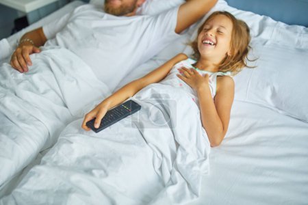 Photo for Father and his young daughter enjoy a restful sleep side by side in a cozy, sunlit bedroom, holding a television remote control, Morning Hours - Royalty Free Image