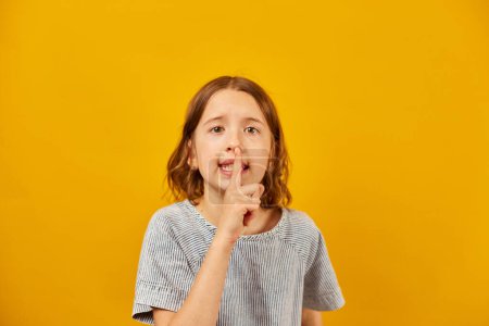 Téléchargez les photos : Une jeune fille se tient devant une toile de fond jaune vif, plaçant son index sur ses lèvres dans un geste universel pour le calme ou le secret. - en image libre de droit