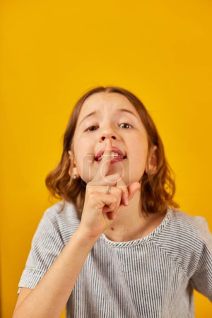 Photo for A young girl stands before a vivid yellow backdrop, placing her index finger over her lips in a universal gesture for quiet or secrecy. - Royalty Free Image