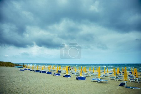 Empty beach chairs and umbrellas await visitors on a sandy shore, as a tempestuous sky looms overhead.