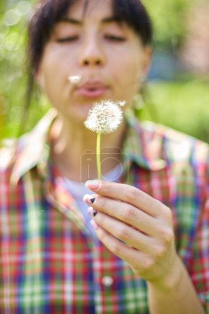 A woman gently blows on a dandelion, scattering seeds in a vibrant garden, Springtime