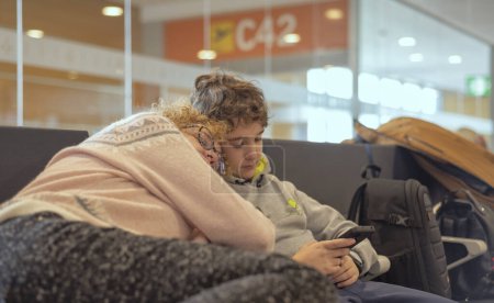 Photo for A tired mom sleeping in an airport on her teenage son's shoulder - Royalty Free Image