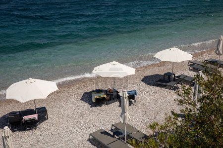 White umbrellas, sunbeds on sundy pebble beach with turquoise crystal clear water in Tyros town, Peloponnese, Argolic Gulf, Myrtoan Sea, GREECE in summer morning. Horizontal.