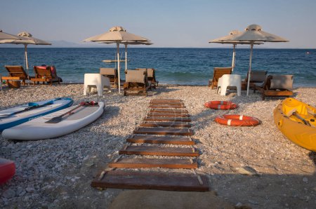 Path to the beach with umbrellas, sunbeds on sundy pebble beach and turquoise crystal clear water. Summer season sea vacation concept. Horizontal.