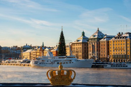 Téléchargez les photos : Le symbole de la couronne royale suédoise, de couleur or, devant Stockholm skyline en hiver, avec l'arbre de Noël dans la vieille ville en arrière-plan. - en image libre de droit