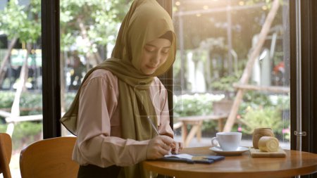 Téléchargez les photos : Une femme musulmane asiatique à la mobilité ascendante profitant d'un moment de détente dans le café par une journée ensoleillée - en image libre de droit