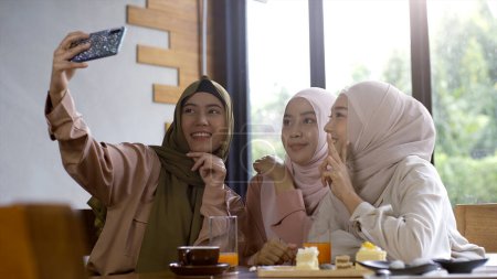 Téléchargez les photos : Un groupe d'amis musulmans asiatiques réussissant vers le haut mobiles savourer un café tranquille rassemblement par une journée ensoleillée lumineuse et joyeuse - en image libre de droit
