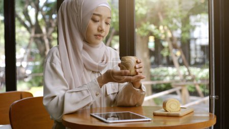 Téléchargez les photos : Une femme musulmane asiatique à la mobilité ascendante profitant d'un moment de détente dans le café par une journée ensoleillée - en image libre de droit