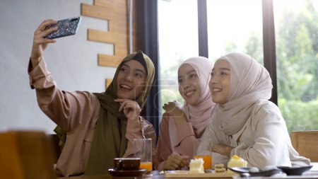 Téléchargez les photos : Un groupe d'amis musulmans asiatiques réussissant vers le haut mobiles savourer un café tranquille rassemblement par une journée ensoleillée lumineuse et joyeuse - en image libre de droit