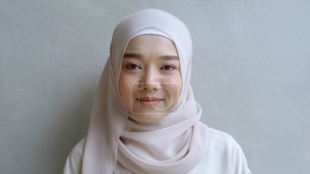 Téléchargez les photos : Gros plan Portrait d'une jeune femme musulmane asiatique vêtue du traditionnel hijab regardant avec confiance et souriant à la caméra avec le mur blanc comme arrière-plan - en image libre de droit
