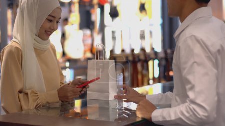 Photo for An upwardly mobile Asian Muslim woman using a mobile phone - smartwatch to pay for a product at a sale terminal with nfc identification payment for verification and authentication - Royalty Free Image