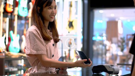 Téléchargez les photos : Jeune femme asiatique utilisant un téléphone mobile pour acheter un produit au terminal du point de vente dans un magasin de détail avec communication en champ proche technologie de paiement d'identification par radiofréquence nfc - en image libre de droit