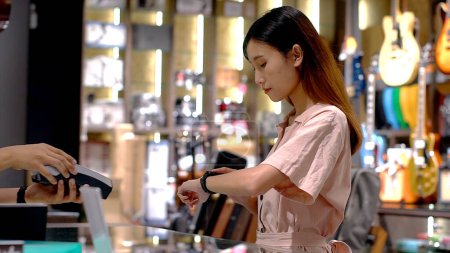 Foto de Mujer asiática joven que usa smartwatch para comprar productos en el terminal de punto de venta en una tienda minorista con tecnología de pago de identificación de radiofrecuencia nfc de comunicación de campo cercano - Imagen libre de derechos