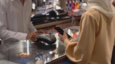 Foto de Una mujer musulmana asiática ascendentemente móvil que usa un teléfono móvil: smartwatch para pagar un producto en una terminal de venta con pago de identificación nfc para verificación y autenticación - Imagen libre de derechos