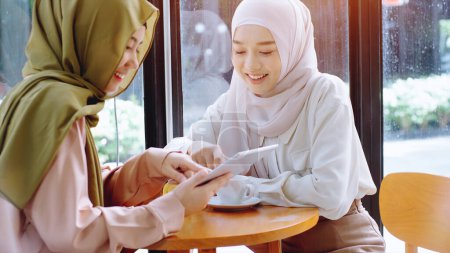 Photo for Young beautiful Asian Muslim women enjoying a relaxing moment working and playing with mobile phone in the coffee shop on a bright sunny day - Royalty Free Image