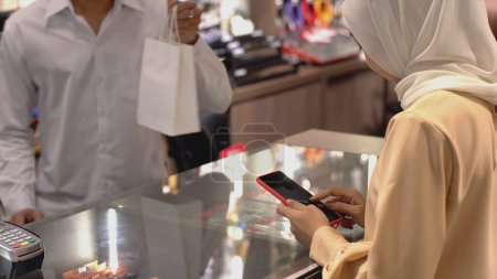 Photo for An upwardly mobile Asian Muslim woman using a mobile phone to pay for a product at a sale terminal with nfc identification payment for verification and authentication - Royalty Free Image