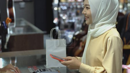 Photo for An upwardly mobile Asian Muslim woman using a mobile phone to pay for a product at a sale terminal with nfc identification payment for verification and authentication - Royalty Free Image