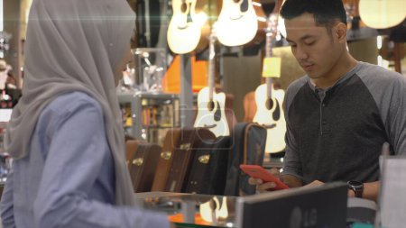 Téléchargez les photos : Un homme musulman asiatique à la hausse utilisant un téléphone portable - smartwatch pour payer un produit dans un terminal de vente avec paiement d'identification nfc pour vérification et authentification - en image libre de droit