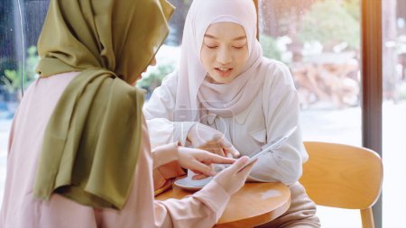 Téléchargez les photos : Jeunes belles femmes musulmanes asiatiques profitant d'un moment de détente en travaillant et en jouant avec un téléphone portable dans le café par une journée ensoleillée - en image libre de droit