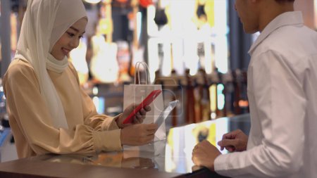 Téléchargez les photos : Une femme musulmane asiatique en mouvement vers le haut utilisant un téléphone portable - smartwatch pour payer un produit dans un terminal de vente avec paiement d'identification nfc pour vérification et authentification - en image libre de droit