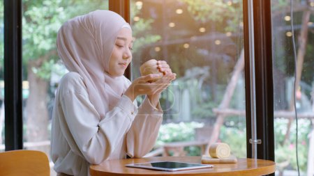 Téléchargez les photos : Jeune belle asiatique musulmane profiter d'un moment de détente travailler et jouer avec appareil mobile dans le café par une journée ensoleillée brillante - en image libre de droit