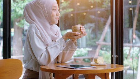 Photo for Young beautiful Asian Muslim woman enjoying a relaxing moment working and playing with mobile phone in the coffee shop on a bright sunny day - Royalty Free Image