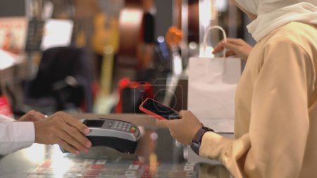 Téléchargez les photos : Une femme musulmane asiatique en mouvement vers le haut utilisant un téléphone portable - smartwatch pour payer un produit dans un terminal de vente avec paiement d'identification nfc pour vérification et authentification - en image libre de droit