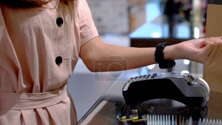 Photo for Young Asian woman using mobile phone - smartwatch to purchase product at the point of sale terminal in a retail store with near field communication nfc radio frequency identification payment - Royalty Free Image
