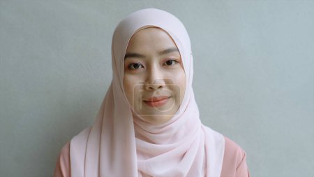 Téléchargez les photos : Gros plan Portrait d'une jeune femme musulmane asiatique vêtue du traditionnel hijab regardant avec confiance et souriant à la caméra avec le mur blanc comme arrière-plan - en image libre de droit