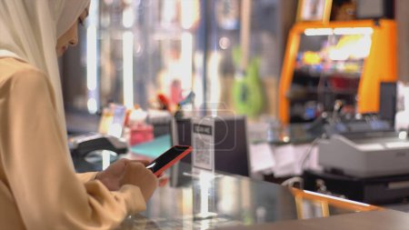 Téléchargez les photos : Un musulman asiatique à la hausse - une femme utilisant un téléphone portable pour scanner le code QR afin de payer au terminal de vente avec paiement d'identification pour vérification et authentification - en image libre de droit