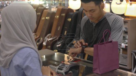 Photo for An upwardly mobile Asian Muslim man using a mobile phone - smartwatch to pay for a product at a sale terminal with nfc identification payment for verification and authentication - Royalty Free Image
