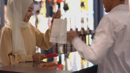 Photo for An upwardly mobile Asian Muslim woman using a mobile phone - smartwatch to pay for a product at a sale terminal with nfc identification payment for verification and authentication - Royalty Free Image