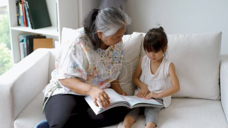 Photo for An elderly Asian woman - Grandmother Teaching Her Granddaughter to read at home on a beautiful morning sunshine through the large living room window - Royalty Free Image