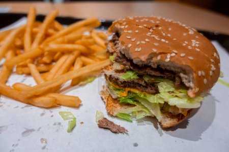 Téléchargez les photos : Gros plan d'un double cheeseburger mordu sur un plateau avec des frites fraîchement cuites dans un restaurant de restauration rapide dans un centre commercial - en image libre de droit