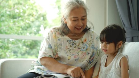 Téléchargez les photos : Une femme asiatique âgée - Grand-mère enseigne à sa petite-fille à lire à la maison sur un beau soleil du matin à travers la grande fenêtre du salon - en image libre de droit