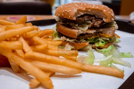 Photo for Close up of a bitten double cheeseburger on a tray with freshly cooked french fries inside a fast food restaurant in shopping mall - Royalty Free Image