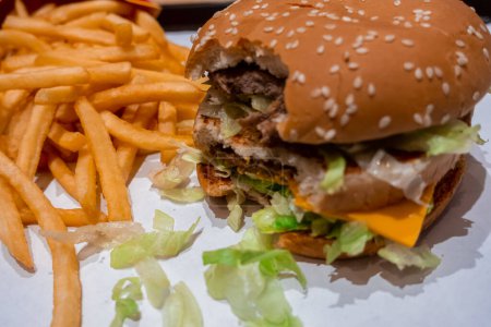 Foto de Primer plano de una hamburguesa doble de queso mordida en una bandeja con papas fritas recién cocinadas dentro de un restaurante de comida rápida en el centro comercial - Imagen libre de derechos