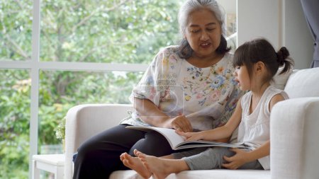Téléchargez les photos : Une femme asiatique âgée - Grand-mère enseigne à sa petite-fille à lire à la maison sur un beau soleil du matin à travers la grande fenêtre du salon - en image libre de droit