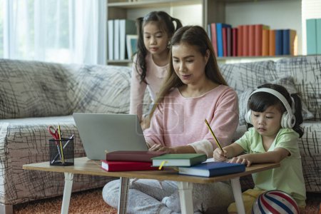 Foto de Una hermosa madre asiática ocupada trabajando mientras cuida y enseña a sus hijos en una computadora portátil en casa - Imagen libre de derechos