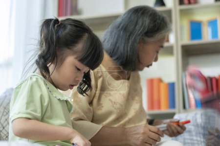 Téléchargez les photos : Une gentille et douce grand-mère asiatique apprenant à sa petite-fille à lire, écrire et dessiner sur un tableau blanc à la maison - en image libre de droit