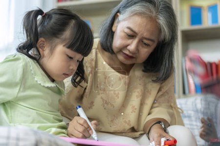 Téléchargez les photos : Une gentille et douce grand-mère asiatique apprenant à sa petite-fille à lire, écrire et dessiner sur un tableau blanc à la maison - en image libre de droit