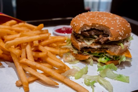 Photo for Close up of a bitten double cheeseburger on a tray with freshly cooked french fries inside a fast food restaurant in shopping mall - Royalty Free Image