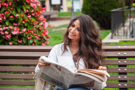 Téléchargez les photos : Belle, jeune femme dans le parc lecture presse avec des sacs à provisions - en image libre de droit